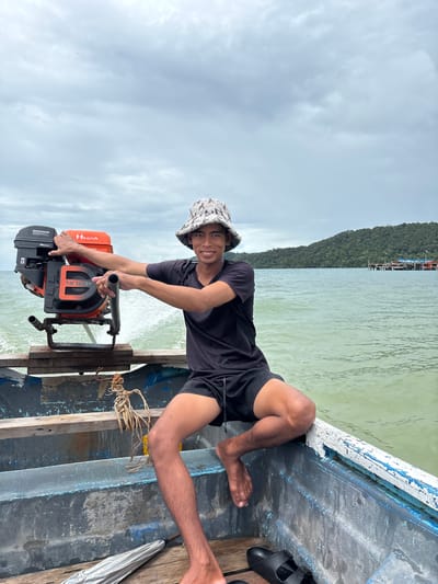 A young man wearing a bucket hat and black t-shirt and shorts is posing for a photo in a boat. He is sitting with his legs bent and is holding onto a small outboard motor attached to the side of the boat. The motor is orange and black with "18.0 HP" and "HONDA" visible on it. He is smiling at the camera. The boat appears to be on water with a green hue, suggesting it might be a river or coastal area. In the background, a tree-covered hill and some buildings on stilts can be seen. The sky is cloudy. The scene suggests a casual outdoor activity, likely in a tropical or coastal region, given the presence of the boat, engine, and lush vegetation. The photo appears to be taken during the daytime under overcast skies.