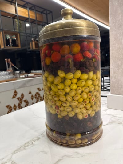 A large, clear cylindrical vessel with a brass-toned metallic lid stands on a white, grey-veined marble countertop. The vessel is filled with a clear liquid and stratified layers of preserved foodstuffs. From top to bottom, the visible layers include: spherical and ovoid red and orange items, resembling small peppers or cherry tomatoes; a dense layer of dark, shriveled, purple-brown olives or dried fruits; a thick mid-section of pale yellow-green ovoid olives, some exhibiting red pimento stuffing; and a base layer of dark purple-black olives. The immediate background to the left reveals a counter with a geometric brown and white triangular pattern, behind which are dark metal framed shelves holding wooden containers and various blurred glassware and metal objects. The background to the right consists of a plain, light-colored vertical surface. Illumination is provided by overhead artificial light sources. This scene is situated within a commercial establishment in Zeytinburnu, Turkey.
