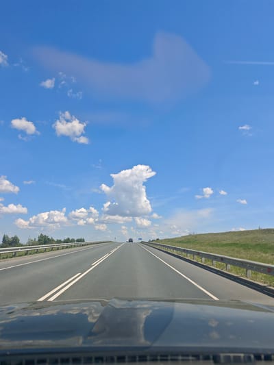 A point-of-view shot from inside a vehicle shows a two-lane asphalt road with white dividing lines, leading into the distance under a bright blue sky with scattered white clouds. The road is bordered on the left by a line of trees and on the right by a grassy embankment topped with a metal guardrail. A dark-colored vehicle is visible in the distance on the road. The hood of the vehicle from which the photo is taken is visible in the foreground, reflecting the sky and clouds.