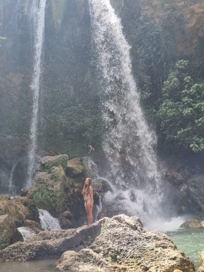 A young woman in a bikini stands on a rock in front of a waterfall. Another person, also in swimwear, is visible in the background, further back and partially obscured by the waterfall. The setting is a natural environment with a large waterfall cascading down a rocky cliff face covered in greenery. The water flows into a pool at the bottom. The image appears to be taken in the town of Anna, Spain, known for its waterfalls and natural beauty.
