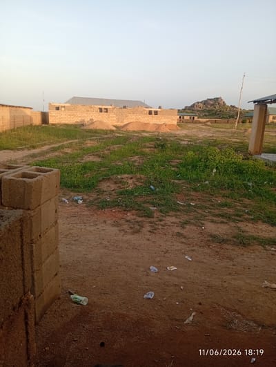 The image depicts an outdoor scene in Du, Nigeria. In the foreground, a stack of concrete blocks is visible on the left. The ground is primarily dirt with patches of dry grass and scattered debris, including plastic bottles and wrappers.
In the mid-ground, unfinished construction is evident. A building made of concrete blocks with an open roof structure is partially constructed, with piles of sand nearby. Further back, several other low-rise buildings with corrugated metal roofs are present.
The background features a prominent rocky hill formation. A single utility pole with wires is visible. The sky is clear and light, suggesting it is either early morning or late afternoon. The overall impression is a rural or semi-developed area undergoing construction. The timestamp "11/06/2026 18:19" is overlaid on the bottom right of the image.