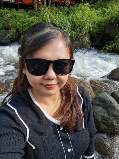 A woman wearing dark sunglasses and a black sweater with white stripes and pearl accents poses in front of a waterfall. Lush green vegetation grows nearby, and the river water cascades over rocks. The woman's hair is brown and layered. Her lips are painted a reddish hue, and she appears to be smiling slightly. The scene appears to be taken outdoors on a sunny day.
