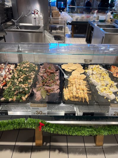 This media file captures a deli or prepared food counter in Rozzano, Italy, likely during the holiday season. The central focus is a refrigerated glass display case showcasing various pre-prepared dishes in black trays.
The visible dishes, from top to bottom, include:
* A white, creamy salad-like dish topped with yellow and dark ingredients, possibly potatoes or corn and herbs.
* Numerous skewers, likely shrimp or chicken, alongside breaded or fried items.
* Slices of roasted meat, such as roast beef, garnished with bay leaves and other seasonings.
* A green salad featuring mixed greens and white protein chunks, possibly chicken or cheese.
* A salad containing red radicchio and other ingredients.
Alongside the food, several white pricing labels are visible. One prominent label states "Menú delle Feste" (Menu of the Festivities/Holidays) with "PRODUZIONE PER" (Production for) and a price of "€30,90". Other clearly visible prices include "€34.90", "€33.90", and a partial "€20". A festive green garland adorned with a small red bow is attached to the side of the display case, reinforcing the holiday theme.
The background reveals a commercial kitchen area with stainless steel countertops, a sink with a faucet, cleaning product bottles, and industrial kitchen equipment. Reflections of people and the indoor environment are faintly visible on the glass and metal surfaces, suggesting customer or staff presence. The scene depicts a bustling food service environment offering ready-to-eat meals, possibly for festive occasions.