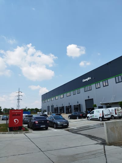 In Cernica, Romania, a sunlit industrial area is depicted, dominated by a large, modern warehouse. The warehouse, with its dark gray facade and a prominent "Homplex" logo in white, features multiple loading bays with green trim. The sky above is a clear blue, punctuated by fluffy white clouds. In the foreground, a concrete lot is partially occupied by parked cars. A red electric charging station with a lightning bolt symbol is positioned near the left edge of the frame, displaying the website "www.zenesis.com". Several vehicles are visible, including a dark blue Dacia Logan with a Romanian license plate "B 260 YNS", a black sedan, and two white vans, one of which is branded with "SIXT". A tall electricity pylon stands in the middle ground, suggesting the presence of power infrastructure. The overall scene is one of an active logistics or industrial facility on a bright day.