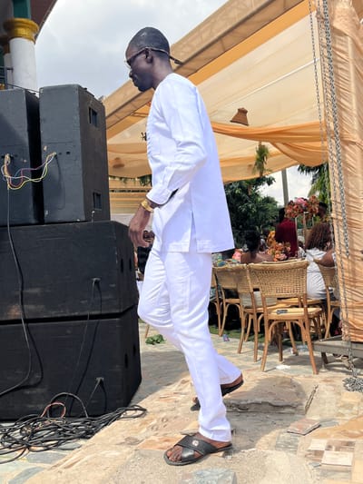 An outdoor event is depicted in Kumasi, Ghana, under a light brown draped canopy. A dark-skinned male subject, of medium build, is positioned mid-stride, walking from the viewer's left to right. He wears a white long-sleeved tunic with side slits, white trousers, and black leather slip-on sandals featuring a gold emblem. He has dark-rimmed glasses, a short hairstyle or tied-back dreadlocks, and a gold-colored wrist cuff on his left wrist. His head is turned slightly to his right.
To the left, two large black professional audio speakers are stacked vertically, with multi-colored audio cables (red, blue, yellow, white) connected at the top unit. A coiled bundle of black cables rests on the ground near the speaker base. In the background, several individuals are seated at round tables with wicker-style chairs. Tables are decorated with floral arrangements in shades of pink, orange, and white. A woman in a red garment is partially visible seated in the background. A woven, bell-shaped decorative item hangs from the canopy structure. Metal chains hang vertically along the right edge of the frame. The ground surface consists of irregular stone paving, with some areas of exposed earth. Green foliage is visible beyond the canopy structure in the distant background, and a white and gold ornate structure is partially visible in the far upper-left corner. The sky is overcast.