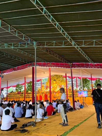 A large group of people are seated on a beige matted floor beneath a white and red tent with a green tarp roof. The people are all wearing white or red clothing. There is a large golden Buddha statue on a stage at the back of the tent, surrounded by flowers. A man wearing a black hat and white shirt is standing in front of the stage holding a camera. A man wearing a black shirt with a black strap across his chest is standing to the right of the image and appears to be taking pictures as well. A TV screen is displaying a video image in the upper right corner of the tent. The photo was taken in Mungkid, Indonesia, at the coordinates -7.605, 110.23.