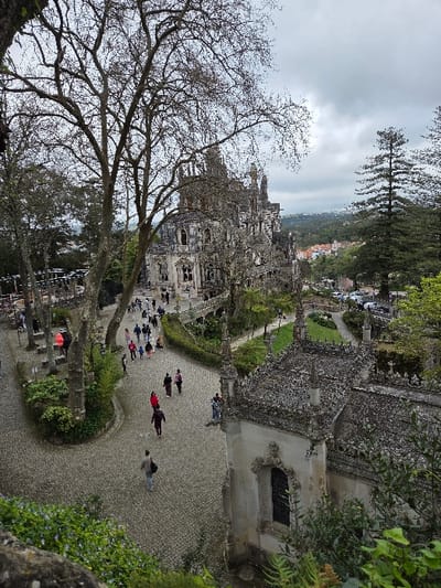 The image captures an elevated view of the grounds of Quinta da Regaleira in Sintra (Santa Maria e São Miguel, São Martinho e São Pedro de Penaferrim), Portugal, under an overcast sky. The central structure is the Palácio da Regaleira, a multi-story building featuring weathered grey stone with intricate Gothic Revival and Romantic architectural details, including numerous spires, turrets, and ornate window frames. A large, deciduous tree with bare branches partially obscures the palace on the left side of the frame.
In the mid-ground, to the lower right, a smaller, ornate stone building, possibly a chapel or annex, is visible. It features a decorative, spired roof and an intricately carved window opening. The foreground and mid-ground are dominated by wide, curved pathways paved with grey cobblestones (*calçada portuguesa*). Approximately 30 to 50 individuals, appearing to be visitors, traverse these paths, moving singly or in small groups. Their clothing varies in color, with some wearing vibrant hues like red and orange, while others opt for darker garments.
Lush green vegetation, including bushes, ground cover, and tall evergreen trees, frames the pathways and structures. In the distant background, beneath a heavily clouded sky, a residential area with red-tiled roofs is visible amidst undulating green hills. A road with several parked cars can be observed in the middle distance on the right.