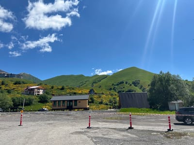 The image depicts a clear, sunny day in Kumlistsikhe, Georgia. In the foreground, a gravel parking lot is marked with red and white striped bollards and caution tape, suggesting a temporary closure or designated area. A dark-colored SUV is parked on the right side of the lot. Behind the parking area, there are several buildings. A small, wooden structure with a grey metal roof sits on stilts, and a larger building with a dark, slanted metal roof is to its right. Further up a lush, green hillside dotted with yellow wildflowers, a multi-story building with a light-colored facade and a reddish-brown roof is visible. Rolling, verdant mountains form the backdrop under a bright blue sky with scattered white clouds and strong sun rays.