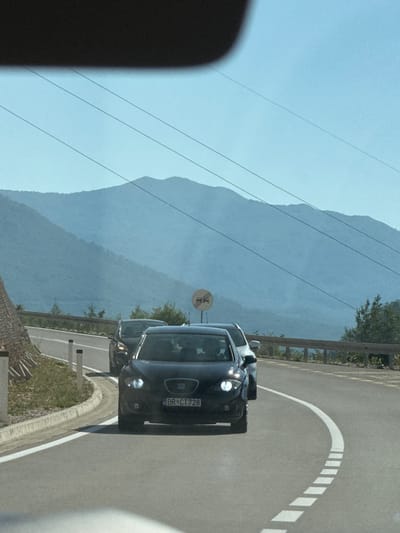 In Šipačno, Montenegro, on a clear day, several cars are seen driving on a paved road that curves to the right. In the foreground, a black car with license plate "BR CT 728" is the closest vehicle. Behind it, another car is visible. The road is marked with white lines, a solid line on the right and dashed lines on the left, indicating lane separation. A traffic sign showing two cars side-by-side, possibly indicating an overtaking restriction or a passing zone, is visible in the background. The surrounding environment includes a grassy embankment on the left side of the road, topped with a wooden fence-like structure. Further in the background, lush green trees and vegetation line the road. In the distance, a mountain range with multiple layers of hazy blue mountains stretches across the horizon under a bright, clear blue sky. Power lines are strung across the sky.