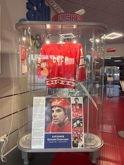 This media file shows a display case containing a red hockey jersey with white stripes, bearing the Cyrillic letters "CCCP". The jersey is prominently displayed in a museum or exhibit, with spotlights illuminating it. Next to the jersey, a large portrait of a hockey player wearing a helmet and jersey is displayed, along with accompanying text. The text includes biographical information about Valery Borisovich Kharlamov, born January 14, 1948, and deceased August 27, 1981. There are also smaller inset photos showing multiple hockey players in action and posed. The background features red flooring with a circular pattern, and large blue block letters spelling out "ЦСКА" (CSKA), a famous Russian sports club. The Olympic rings are also visible behind the jersey. The overall setting appears to be indoors, likely a sports museum or hall of fame, given the artifacts and informational displays. The lighting suggests it is daytime or the exhibit is well-lit. The images and text evoke a sense of sports history and national pride, likely celebrating the achievements of Soviet hockey.