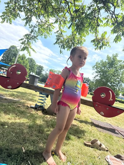 A young girl with blonde hair and blue eyes is posing for the camera outdoors. She is wearing a colorful rainbow swimsuit with a cartoon ice cream graphic and orange arm floaties. The girl is standing on a grassy area, with trees and a blue sky with clouds in the background. In the foreground, there are parts of a red playground structure with metal springs attached. A pair of tan slides are on the grass to the right of the girl. The scene appears to be a sunny day, likely in the afternoon, given the lighting and shadows. The girl has a neutral expression. The word "CE" and a number "#58542" are visible on the arm floatie.