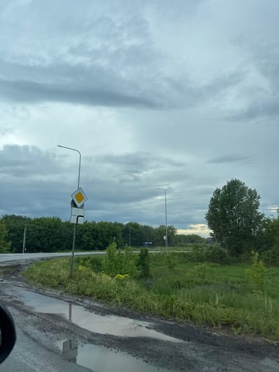 The image captures a roadside scene in Нестюково, Russia, under an overcast sky, suggesting recent rainfall. The perspective appears to be from inside a vehicle, with a dark curve, possibly part of a dashboard or steering wheel, visible in the bottom right corner.
The sky dominates the upper portion of the frame, filled with dense, gray and white clouds that show some streaky patterns, indicative of an unsettled or post-storm weather front. A brighter patch of sky is visible on the horizon towards the right, hinting at possible clearing or the sun beginning to peek through.
Below the sky, a green landscape unfolds. A paved road stretches into the distance, bordered by a wide, unpaved or dirt shoulder. This shoulder is marked by several significant puddles, reflecting the dull sky and indicating that rain has recently fallen. Lush green grass and low bushes line the immediate roadside, with denser foliage and trees forming a treeline further back on both sides of the road. One large, full-crowned tree stands out in the mid-ground on the left.
Several elements of road infrastructure are present. Two prominent street lights with curved arms are visible. Attached to one of these poles are two road signs: a yellow diamond with a white border, which is a European sign for "priority road," and a square sign depicting a black main road turning sharply right with a minor road joining from the left, indicating the priority road's direction at an intersection. Further down the road, a small, circular white sign with the number "40" in black text is faintly visible on a pole, likely a speed limit sign. The overall scene conveys a quiet, rural or semi-rural environment on a cloudy, post-rain day.
