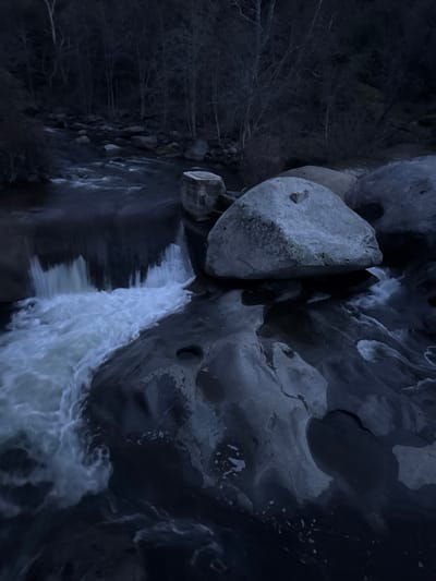 This image depicts a natural river scene in Three Rivers, United States, under low light conditions. The primary subject is a dark, fast-flowing river with sections of rapids and a small waterfall. The water exhibits motion blur, indicating a long exposure, with white foam streaking over dark, eroded bedrock.
Numerous large, grey boulders are present within and alongside the river. A prominent, light-colored, smooth boulder is centrally located, with a small, dark, heart-shaped stone resting on its upper surface. To the left of this central boulder, a rectangular concrete-like block is partially submerged at the edge of the rushing water. The riverbed is composed of dark, irregularly shaped bedrock featuring several smooth, water-filled potholes.
The far riverbank is densely vegetated with bare deciduous trees and shrubs, suggesting a winter or late autumn season. The overall illumination is dim, casting a cool, blue-grey tone over the entire scene. There are no individuals, animals, or discernible human activities depicted.