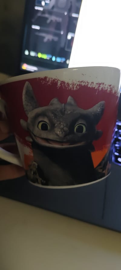 A close-up shot shows a white mug with a red background and a drawing of Toothless, the dragon from "How to Train Your Dragon," on it. The mug is being held by a person's hand, partially visible on the left. In the background, a blurred computer monitor displays a menu with weapon icons and text, suggesting a gaming environment. A portion of a keyboard with illuminated keys is visible on the right. The overall scene is dimly lit, with the primary light source appearing to be from the monitor and an overhead light. The specific location in Skopin, Russia, cannot be determined from the visual information provided.