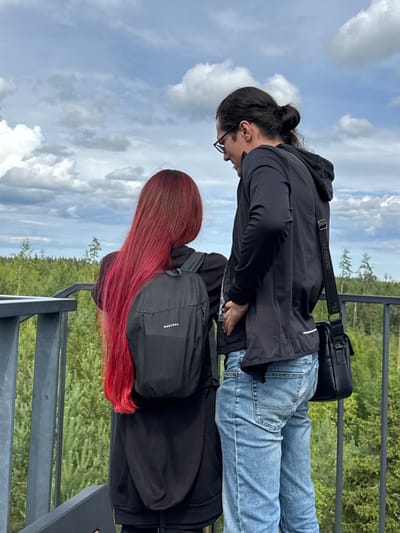 Two individuals, a woman with long red hair and a man with dark hair in a bun, stand on a viewing platform overlooking a dense forest. The woman, facing away from the camera, wears a black hoodie and a black backpack with the "QUECHUA" logo visible. The man, facing towards the woman, wears a black hooded jacket, blue jeans, and glasses. He has a black messenger bag slung across his body. The environment consists of a metal railing in the foreground, a lush green forest with scattered trees in the middle ground, and a cloudy sky with blue patches in the background. The location is identified as Peski, Russia.