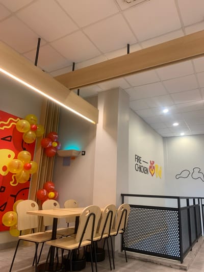The image displays the interior of a "FIRE CHICKEN" restaurant located in Calukan RT 1 RW 5 Keboansikep, Indonesia. The scene is brightly lit with a white grid ceiling featuring recessed spotlights and suspended light-colored wooden beams, one equipped with a linear LED light strip. The walls are painted white. On the left wall, a large red and yellow graphic design is visible, decorated with multiple clusters of red and yellow balloons, some translucent and bearing an indistinct logo. A light-colored wooden wall-mounted fixture below the balloons emits an upward blue light. In the foreground, two light-colored wooden rectangular tables with black metal bases are arranged. Six matching wooden chairs with black metal frames and circular cutouts on their backrests are positioned around one of the tables. A white structural column stands centrally. The right wall features black lettering spelling "FIRE - CHICKEN," accompanied by stylized red and yellow chicken wing graphics, followed by two outlined white chicken wing shapes. A black metal railing with perforated panels delineates a change in floor level on the right side of the frame. No people, actions, or interactions are present in the image.