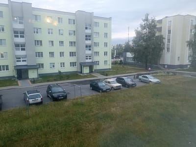 The image depicts an exterior view of an apartment building in Mikashevichy, Belarus. The building is light green with gray accents, featuring numerous windows. In front of the building, a paved area serves as a parking lot with several cars parked. Adjacent to the parking lot is a grassy area. A person is visible walking near the apartment building on the right side of the image. The sky is overcast.