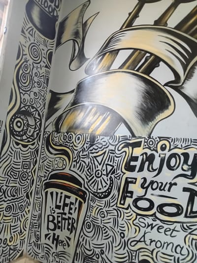 The media file displays a vibrant, two-part mural painted on a wall or ceiling, featuring contrasting artistic styles. The artwork is predominantly in black, white/light grey, and metallic yellow/gold tones.
On the left side of the image, large, flowing, and shaded ribbon-like or sail-like elements are depicted. These elements have a three-dimensional quality, rendered with dark shadows and bright yellow/gold highlights, suggesting a glossy or metallic texture. They appear to be part of a larger, abstract structure, possibly resembling ship rigging or decorative scrolls.
The right side of the mural, and a section along the bottom, is covered in dense, intricate doodle art or graffiti-style patterns. These patterns consist of swirling lines, geometric shapes, spirals, and abstract motifs, all outlined in black on a light background, with occasional yellow accents filling in certain shapes or outlines.
Visible text within the doodle section includes "Enjoy your FOOD" rendered in large, stylized black letters with yellow outlines. Below this, "Sweet Aroma" is written in a smaller, script-like font. Further down, integrated into the patterns, is a stylized coffee cup graphic containing the text "LIFE is BETTER <3 Here" in blocky black letters.
The mural creates a lively and dynamic aesthetic, combining bold, illustrative elements with detailed, abstract patterns. The presence of food and aroma-related text, along with the coffee cup, suggests this artwork likely adorns the interior of a cafe, restaurant, or similar establishment in Barako, Nigeria.