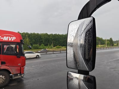 This image captures a view from the driver's seat of a red truck, likely a semi-trailer, parked on a wet asphalt road. The truck's side mirrors are prominent, covered in raindrops, reflecting the surrounding environment. In the foreground, the front left wheel and lower portion of the red truck are visible. A white sedan is seen driving on the road to the left, in the middle ground, with a blurred background of green trees lining the road and a cloudy sky overhead. The scene appears to be on a highway or main road, possibly near the village of Turlaovo, Russia, given the presence of a white sedan and the type of vegetation. The overall atmosphere suggests a damp, possibly rainy day.