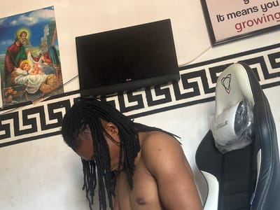 An indoor scene in Ogwashi-Uku, Nigeria, shows a man resting on a light-colored floor with a black and white Greek key pattern rug. The man, with dark skin and long dreadlocks, is lying on his back, shirtless, with his head towards the right side of the frame. His right ear and a portion of his chest are visible.

To his left, part of a black and white racing-style chair is visible. On the chair, a white slipper or slide shoe, still wrapped in clear plastic, is placed. The slipper has a red and blue triangle logo and the partial text "AKWLE" is discernable. Below the man's head, a black LG monitor or tablet is positioned on the floor. To the right of the monitor is a religious picture, depicting figures resembling Mary, Joseph, and baby Jesus in a Nativity-like scene, with a blue sky background. In the lower-left corner, a framed item with upside-down text is visible, reading "growing if it means you" before being cut off or obscured. The overall impression is one of a relaxed, informal setting.