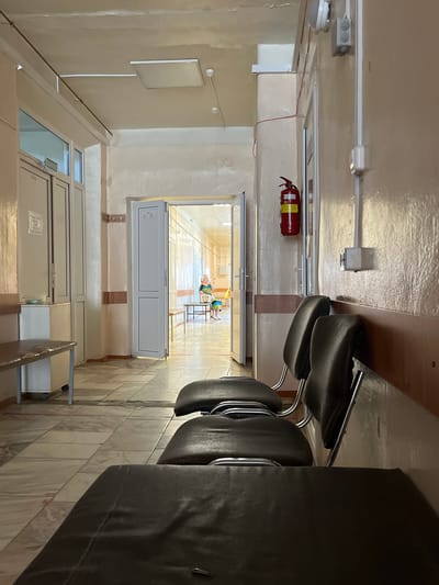 The image shows a hospital corridor in Ikryanoye, Russia. In the foreground, there are several dark leather chairs arranged in a row. To the left, there is a long, narrow table with a white cabinet underneath. Further down the corridor, double doors are open, revealing another section of the hallway. In this area, a woman in a colorful dress is seated on a bench, looking towards the right. A fire extinguisher is mounted on the wall to the right of the open doors. The corridor walls are painted a light pink color, and the floor is tiled with a marble-like pattern. The ceiling has fluorescent lighting fixtures.