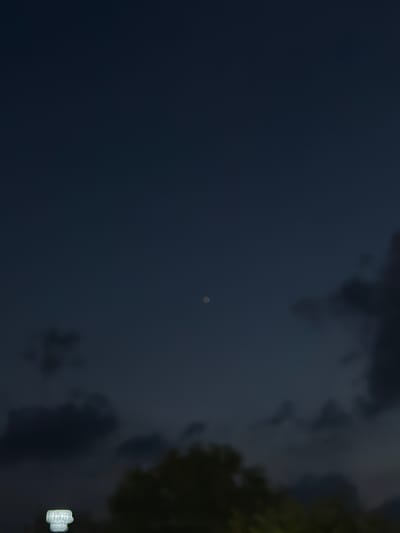The image captures a dark, twilight sky over Uijeongbu-si, South Korea, with a single, small, bright point, likely a planet or star, visible near the center. The lower portion of the frame is occupied by dark, silhouetted clouds and foliage. In the bottom left corner, a blurred, illuminated sign with white text is partially visible. The overall atmosphere is dim and moody, suggesting the transition from day to night.
