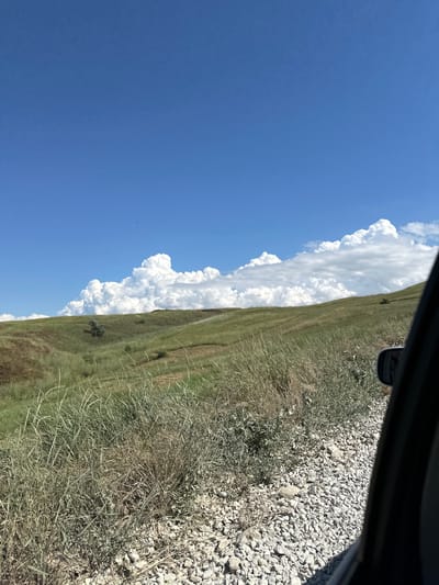This media file depicts a scenic view from a vehicle, looking out over a rolling grassy hillside under a bright blue sky with large, fluffy white clouds. The perspective suggests the photo was taken from inside a car, with the top edge of the frame showing a dark car interior element, possibly the dashboard or window frame, and a portion of a side-view mirror. The ground beside the road is covered in gravel and dry grass. The hillside itself is a mix of green and drier, straw-colored grass, with a few scattered bushes or small trees. The dramatic clouds are a prominent feature, creating a striking contrast with the clear blue sky. The overall impression is of a sunny day in a rural or natural landscape, likely a vast open space like a steppe or prairie. The orientation of the image suggests it was captured while traveling, possibly on a road trip. No people or specific activities are visible, but the setting evokes a sense of journey and natural beauty. The time of day appears to be daytime, given the bright sunlight and clear skies. There is no visible text in the image.