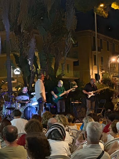 This is an outdoor evening scene in Ospedaletti, Italy, likely on a street or a small piazza, with a band performing. A group of musicians, consisting of a drummer, two guitarists, and possibly a singer, are on a raised platform. The drummer is seated behind a drum kit, one guitarist is playing a green electric guitar, and another is playing a white electric guitar. A singer, wearing a light-colored shirt, stands center stage. In the foreground, an audience is seated in chairs, facing the stage, with their backs to the camera. The background shows a building with balconies and windows, illuminated by streetlights and lights from the performance. Palm trees and other foliage are visible, adding to the Mediterranean atmosphere. The overall impression is of a casual, local musical event.