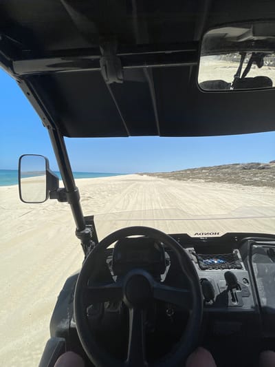 The image shows the interior of a Honda UTV vehicle driving on a beach in La Capilla, Mexico. The vehicle has a black steering wheel, side mirrors, and a roof. Outside, tire tracks are visible in the sand. The ocean is to the left, and vegetation can be seen along the beach. The sky is blue.