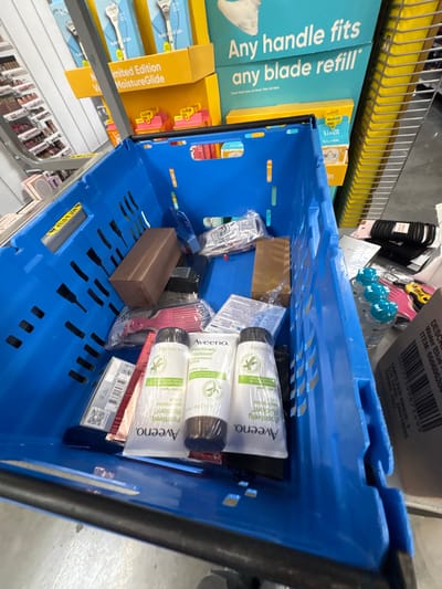 The image depicts the interior of a retail store, located in Tigard, United States. A blue, rigid plastic shopping cart or tote, seen from a high-angle perspective, occupies the majority of the frame. The cart is filled with assorted consumer goods. Prominently displayed in the front of the cart are three identically packaged, shrink-wrapped tubes of "Aveeno Positively Radiant" daily moisturizer. Other contents include a rectangular brown cardboard box, a clear plastic bag containing white items (possibly medical supplies), a smaller brown paper bag, and a partially visible pink plastic hair comb.
In the background to the left, a silver-colored retail display unit with multiple shelves holds various cosmetic products. Centered in the background are large blue and yellow cardboard display boxes for razor blade products, featuring text such as "Any handle fits any blade refill" and "Limited Edition MoistureGlide," suggesting Gillette Venus brand items. To the far right, a vertical yellow metal retail shelving unit is visible. The floor is light grey, characteristic of a commercial retail environment. No individuals are present in the scene, and no specific actions or interactions are depicted beyond the static arrangement of the products within the cart.