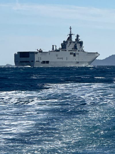 A large, grey naval vessel, possibly a helicopter carrier or amphibious assault ship, sails on a choppy, deep blue sea under a hazy, pale blue sky. The ship has a long, flat deck at the stern and a raised superstructure at the bow, with antennas and what appears to be a helicopter on the stern deck. The vessel is positioned in the middle ground, with the turbulent water dominating the foreground and a hazy, green-covered hill or island in the far right background. The image is taken from a low vantage point, likely from another boat. The scene is not identifiable as being located in Bormes-les-Mimosas, France.