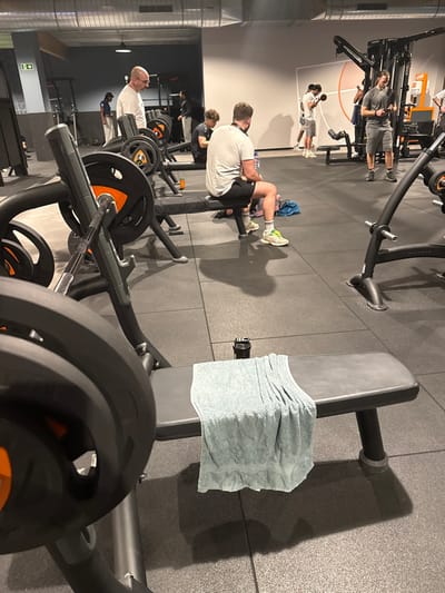The image captures an indoor gym environment, brightly lit by artificial lights, with a dark rubber tiled floor and exposed ductwork overhead. The scene is populated by several individuals, primarily men, engaged in various weight training activities.
In the mid-ground, a man wearing a white t-shirt and dark shorts sits on a grey weight bench, facing away from the camera. His legs are bent, and he wears light-colored athletic shoes. Nearby, a blue fabric item rests on the floor. To the right, another grey weight bench is unoccupied, with a neatly folded light grey towel placed on its seat.
Further back and to the left, another man in a grey t-shirt and shorts stands near a multi-station cable machine, with the brand "MATRIX" faintly visible on some equipment parts. Beside him, a younger individual in a white t-shirt and light shorts appears to be performing an exercise. To the far left, partially obscured, an older man in a white t-shirt is positioned near a barbell rack, which holds numerous weight plates, some featuring orange accents. Other gym-goers are visible in the background, suggesting a moderately busy gym floor.
The activity depicted is typical for a fitness center, with people either actively working out or resting between sets. Shadows from the equipment and individuals are cast on the dark flooring. On the far left wall, a small white sign with an arrow and an unreadable symbol or text, possibly an exit indicator, is visible. Below it, a partial logo or text, potentially related to the gym's brand, can be seen on the wall.
