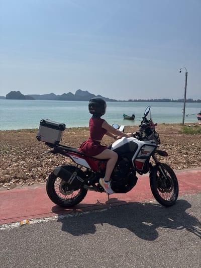 A person wearing a helmet, a maroon top, and maroon shorts sits on a Yamaha motorcycle. The motorcycle is parked next to a road and near a body of water. Mountains are visible in the distance. The motorcycle has a storage container attached to the back. The sky is clear, and there is a shadow cast by the motorcycle and the person.