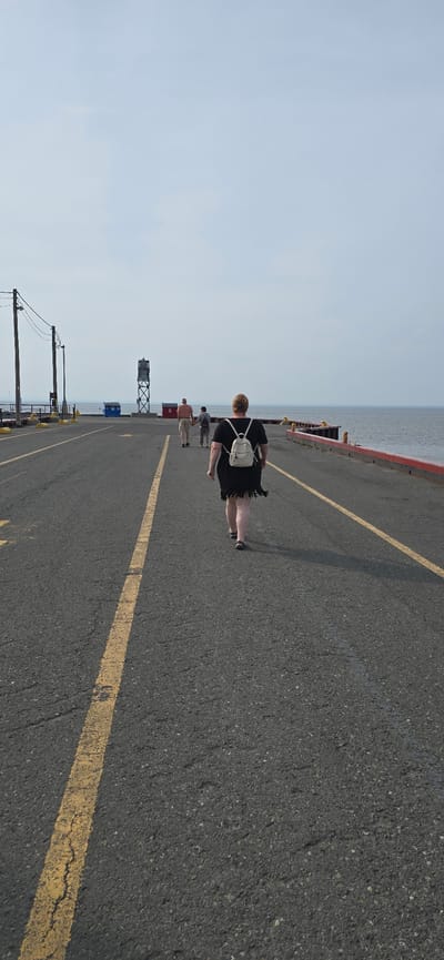 The image depicts a long, paved walkway or pier extending towards the water. A person wearing a black dress and a white backpack is walking away from the camera down the center of the walkway. Further ahead, two other figures, appearing to be an adult and a child, are walking in the same direction. On the left side of the walkway, there are telephone poles with wires. In the distance, a tall structure resembling a lighthouse or navigation tower stands between two small buildings, one red and one blue. The walkway is bordered on the right by a red railing overlooking the water. The sky is overcast. The location is identified as the docks in the city of Trois-Pistoles, Canada.