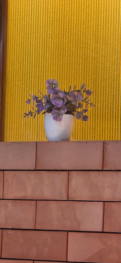 A white ceramic pot containing a faux purple floral arrangement sits on a brick ledge. The pot is centered horizontally and positioned slightly above the midpoint of the image vertically. Behind the pot and floral arrangement is a textured, bright yellow background with vertical striations. The brick ledge extends across the bottom half of the image, with visible grout lines between the reddish-brown bricks. The scene is indoors, likely a home or business, in Urupskiy, Russia. No people are present.