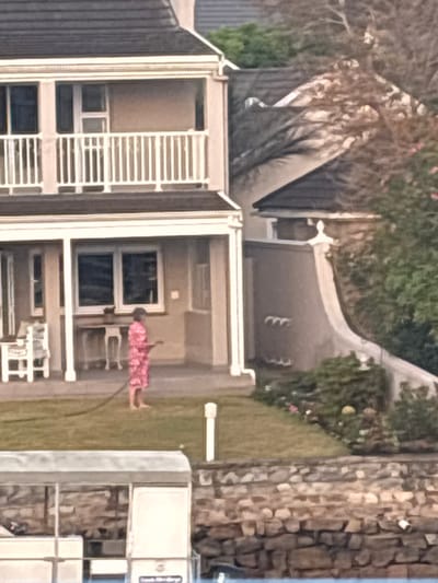 A two-story building with white trim and a gray roof stands adjacent to a green lawn where a person wearing a pink floral patterned outfit uses a garden hose. An area with seating and a table is located under the building's balcony. A tree is to the left of the building and a stone wall is to the right. A sidewalk can be seen behind the structure.