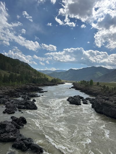 The image depicts a fast-flowing river in the Edigan, Russia area, bordered by black, rocky shores on the right side of the frame. Mountains are visible in the background, layered in shades of green and grey. A clear blue sky, filled with fluffy white clouds, occupies the left side of the frame. The light suggests it's daytime.