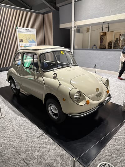 The image shows a cream-colored vintage Subaru 360 car on display in what appears to be a museum or exhibition space in Sumida, Japan. The car is positioned on a black platform over grey herringbone carpet flooring. A descriptive panel in Japanese and English is visible on the wall to the left of the car. The surrounding environment features neutral-toned walls and interior architecture. A person's legs are visible in the background. A security cord is visible around the base of the black platform. The car's interior is partially visible through its windows.