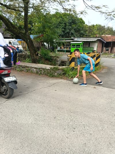 A daytime photograph taken at eye-level depicts a street scene in Dayeuhkolot, Indonesia. In the foreground to mid-ground, a young male, dressed in a light blue t-shirt, blue shorts, and blue athletic shoes, is actively engaged with a light-colored soccer ball on an asphalt road. He is crouched forward, right hand on the ball, with his head turned towards the left. Behind him, yellow and black striped road barriers border a concrete curb and an area of overgrown grass and small trees. To the far left, the rear section of a red motorcycle with a visible license plate "D 6763 ACA" is parked or in transit, with a partial view of a person wearing a white garment and a black backpack. The background features a cluster of simple, utilitarian structures, including a prominent green-fronted stall or shop, all situated beneath a dense canopy of green trees and a partially visible light blue sky. The asphalt road shows minor cracks and surface irregularities.