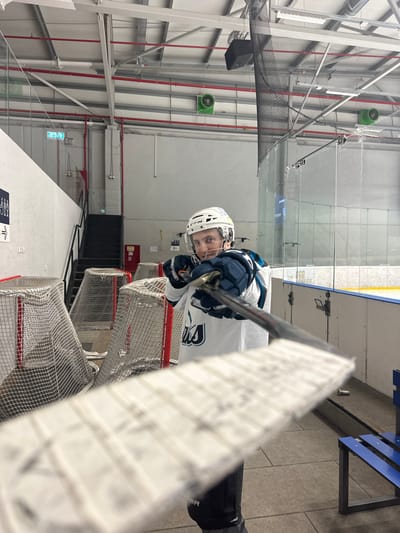 A hockey player in a white helmet and jersey holds a stick, looking towards the camera. He is in an ice rink with multiple hockey nets stacked in the foreground, partially obstructing the view. An ice rink wall and railing are visible in the background. Overhead structures, including piping and a speaker, can be seen on the left. The floor is visible on the right, with a portion of a blue bench.