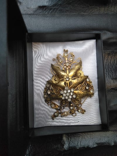 The image displays an ornate, gold-toned metal brooch, studded with numerous clear, faceted stones, presented within an open, rectangular black box. The brooch's design incorporates stylized leaf elements, intricate scrollwork, and a central cluster of stones, with additional stone-adorned dangling components at its base. It rests on a white, rippled, satin-like fabric insert within the box. The exterior of the black box is situated on a dark, textured surface. This scene is observed in Косшы, Kazakhstan.