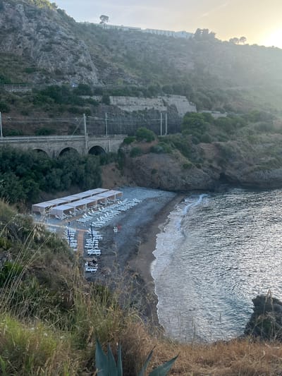 This image depicts a coastal cove in Sperone, Italy, viewed from an elevated perspective overlooking a pebble beach. The scene is illuminated by soft, hazy sunlight, suggesting either early morning or late afternoon.
In the upper portion, a steep, rocky mountainside dominates, covered with sparse green and brown vegetation. A winding road or path is visible traversing the slope, with a small, light-colored vehicle present on it. Higher up, a white building complex, likely a hotel or resort, is nestled among trees on the distant hilltop.
Mid-ground features a railway line running horizontally across the image, supported by overhead electrical infrastructure. Below the railway, a multi-arched stone viaduct spans a section of the terrain, partially obscured by dense green foliage.
The focus shifts to the beach in the lower-mid section, consisting of dark grey pebbles. Twelve rectangular sunshade structures, or pergolas with light-colored fabric tops, are arranged in two parallel rows. Numerous white plastic sun loungers are positioned beneath and around these pergolas; most appear unoccupied. A wooden boardwalk or pathway extends from the foreground onto the beach, leading towards the sunshades. One individual is discernible sitting on a sun lounger near the end of the boardwalk, partially hidden by foreground foliage. A red lifebuoy is mounted on a stand to the right of the loungers.
To the right, the calm, dark water of the sea laps the pebble shore with small white waves. The water surface reflects the overhead light, creating a shimmering effect. A rocky outcrop defines the right edge of the cove where it meets the sea.
The immediate foreground consists of dry, tall grasses and an agave-like plant, framing the panoramic view of the beach and sea below.