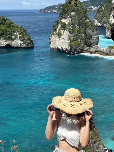 A woman in a white blouse and straw hat stands on a cliff overlooking the sea near Pejukutan, Indonesia. The turquoise water surrounds rock formations covered with vegetation. The sky is partly visible in the distance.