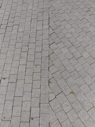 The image shows a close-up, overhead view of a grey paved surface made of interlocking square concrete blocks. The paving is divided by a slightly uneven seam running diagonally from the top left to the bottom right of the frame, suggesting two sections of pavement or a change in pattern. The blocks are laid in a staggered pattern, with slight variations in color and texture, and some minor imperfections or wear are visible, including small cracks and a few stray leaves and debris in the crevices. The overall impression is of an outdoor public walkway, likely a sidewalk or plaza. This scene is consistent with many paved areas found in the city of Târgoviște, Romania.