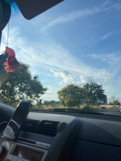 The image is taken from the interior of a car, looking out through the windshield. A pink fuzzy charm hangs from the rearview mirror. The dashboard is visible in the foreground, with a smartphone mounted on a vent. The car's radio display shows "19:21" and "99.1". Outside the car, there are trees, a building, and a clear blue sky with wispy clouds. The location is identified as Graf Ignatievo, Bulgaria.