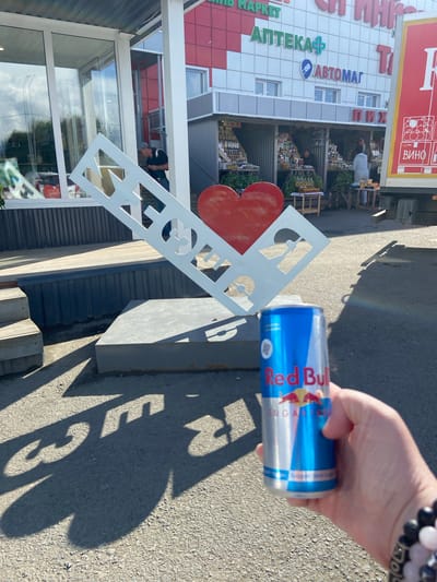 In Inzer, Russia, a person's hand holds a Red Bull can in the foreground. Behind the hand, a large white sculpture shaped like a heart and the letters "МЫ" (We) is visible. The sculpture casts a shadow on the asphalt ground. In the background, there are market stalls selling goods, a building with signs for "АПТЕКА" (Pharmacy) and "АВТОМАТ" (Vending Machine), and a truck. A man is seen walking down stairs in the background.