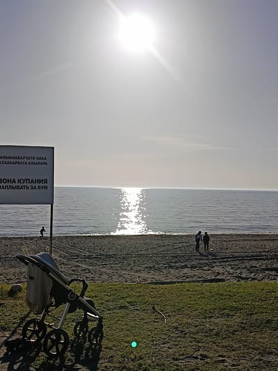 The image shows a beach in Gulrypsh, Abkhazia on a sunny day. In the foreground is a baby stroller parked on a grassy area bordering the beach. Behind the stroller, there is a sandy beach with a sign on the left that appears to be in Russian and another language. In the distance, there are two people walking on the beach, and another person is near the water. The sea extends to the horizon where it meets the sky. The sun is high in the sky, causing a reflection on the water.