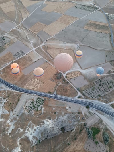 The aerial view captures a landscape that is characteristic of Cappadocia, near Avanos, Turkey. The terrain is a mosaic of agricultural fields, some fallow and others showing patterns of cultivation. A winding paved road runs through the scene, with a few white vehicles visible on it. Scattered across the landscape are several hot air balloons, appearing to be in the process of a flight. One large, pale orange balloon dominates the center, while other smaller balloons, some yellow with red and blue stripes and one light blue with a checkered pattern, are also visible. The ground shows signs of erosion and geological formations, with rocky outcrops and valleys. The time of day appears to be early morning or late afternoon, given the soft lighting.