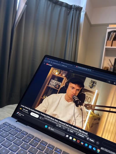 A laptop screen displays a YouTube video titled "Leeds United Eye Gudmundsson, 'Pushing' £35M Muniz Deal, Cresswell Sell-On Revealed (Big Move!)". The video features a man wearing headphones and speaking into a microphone. Behind him is a sign reading "ELLAND ROAD" and the number "10". The YouTube channel is "One Leeds" with 45.2K subscribers. Buttons for subscribe, share, and download are visible, alongside a search bar. The laptop's keyboard is partially visible in the lower right corner.