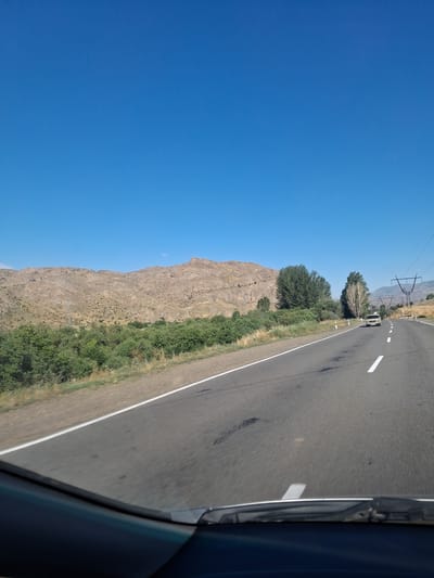 The image depicts a scenic view from a vehicle traveling on a road. The sky is a clear, bright blue, suggesting a sunny day. To the left of the road, a large, rocky mountain rises, its rugged texture and arid appearance indicating a dry climate. A strip of green vegetation, including trees and bushes, grows along the base of the mountain and slopes down towards the road.

The road itself is paved with asphalt and has a white line marking the lane. A single white car is visible on the road, traveling in the same direction as the viewer. In the distance, power lines and poles are visible against the horizon, hinting at human infrastructure in the otherwise natural landscape. The perspective suggests the photo was taken from the passenger seat of a car, with a portion of the car's interior, including a windshield wiper, visible in the foreground on the right side of the image.

The overall setting appears to be a rural or mountainous region, with the location context suggesting Malishka, Armenia. The bright sunlight and clear sky suggest the time of day is likely midday or early afternoon. The scene evokes a sense of travel and exploration through a natural landscape. There is no visible text in the image.